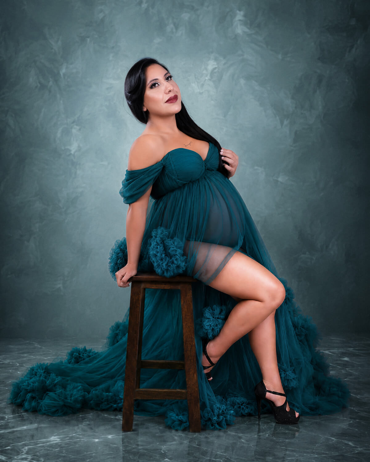 Fotografía artística de embarazo en Cochabamba, Bolivia, de una mujer embarazada posando en un taburete con un vestido de gala verde esmeralda y tul, realizada por 53 Fotografía.