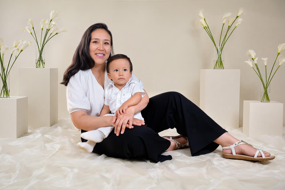 Retrato de estudio de una madre y su hijo pequeño sentados, composición minimalista con flores blancas de fondo. Sesión de fotografía familiar en Cochabamba.