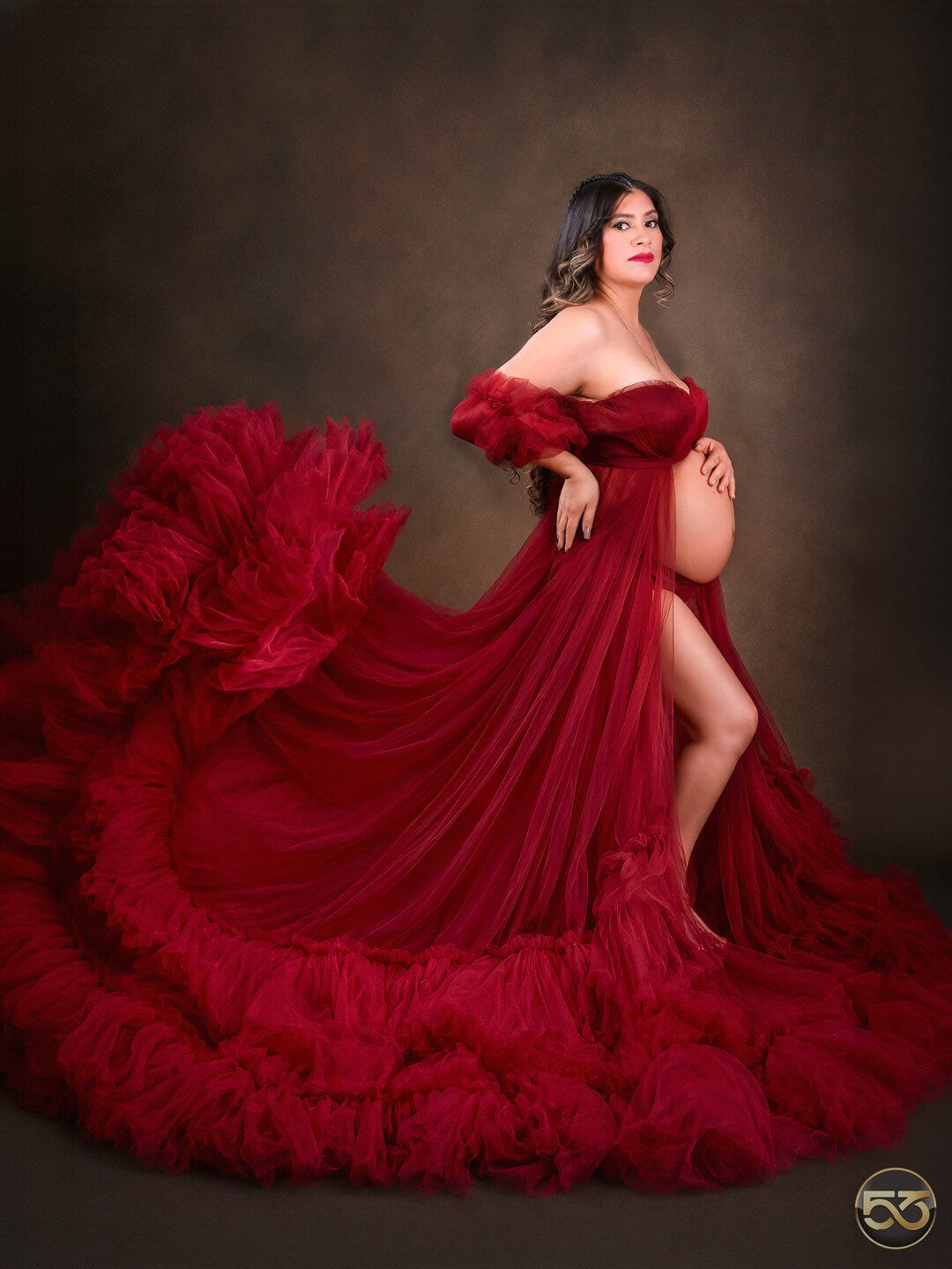 Mujer embarazada con vestido rojo de tul, vientre descubierto y una larga cola con volantes sobre un fondo marrón oscuro.